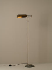 A modern floor lamp with a brass-finished pole and adjustable rectangular shade, mounted on a round dark stone base, photographed against a plain warm beige wall.