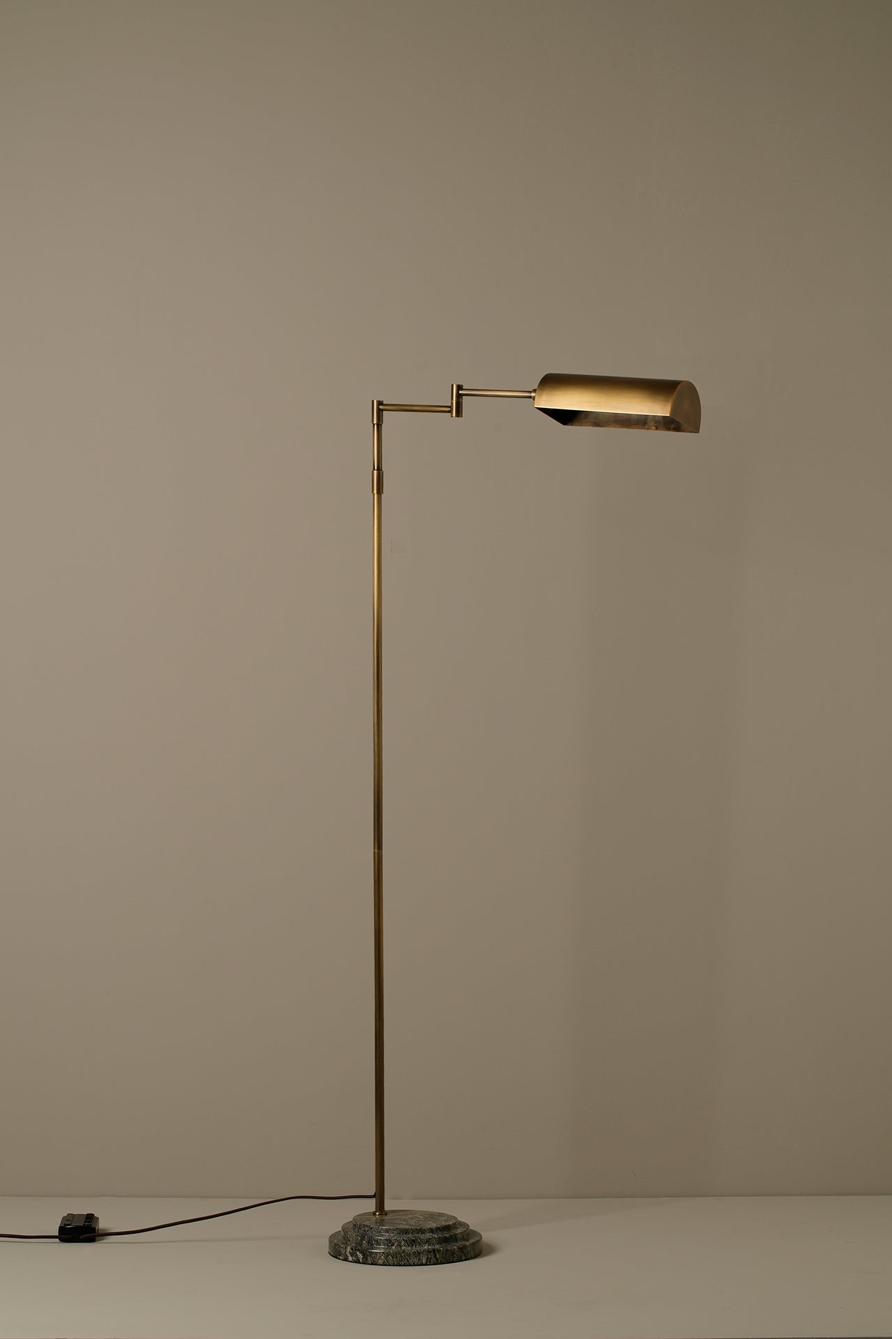 A mid-century modern floor lamp with a slim brass-colored metal stand and a rectangular shade, set on a round stone base against a plain beige wall.