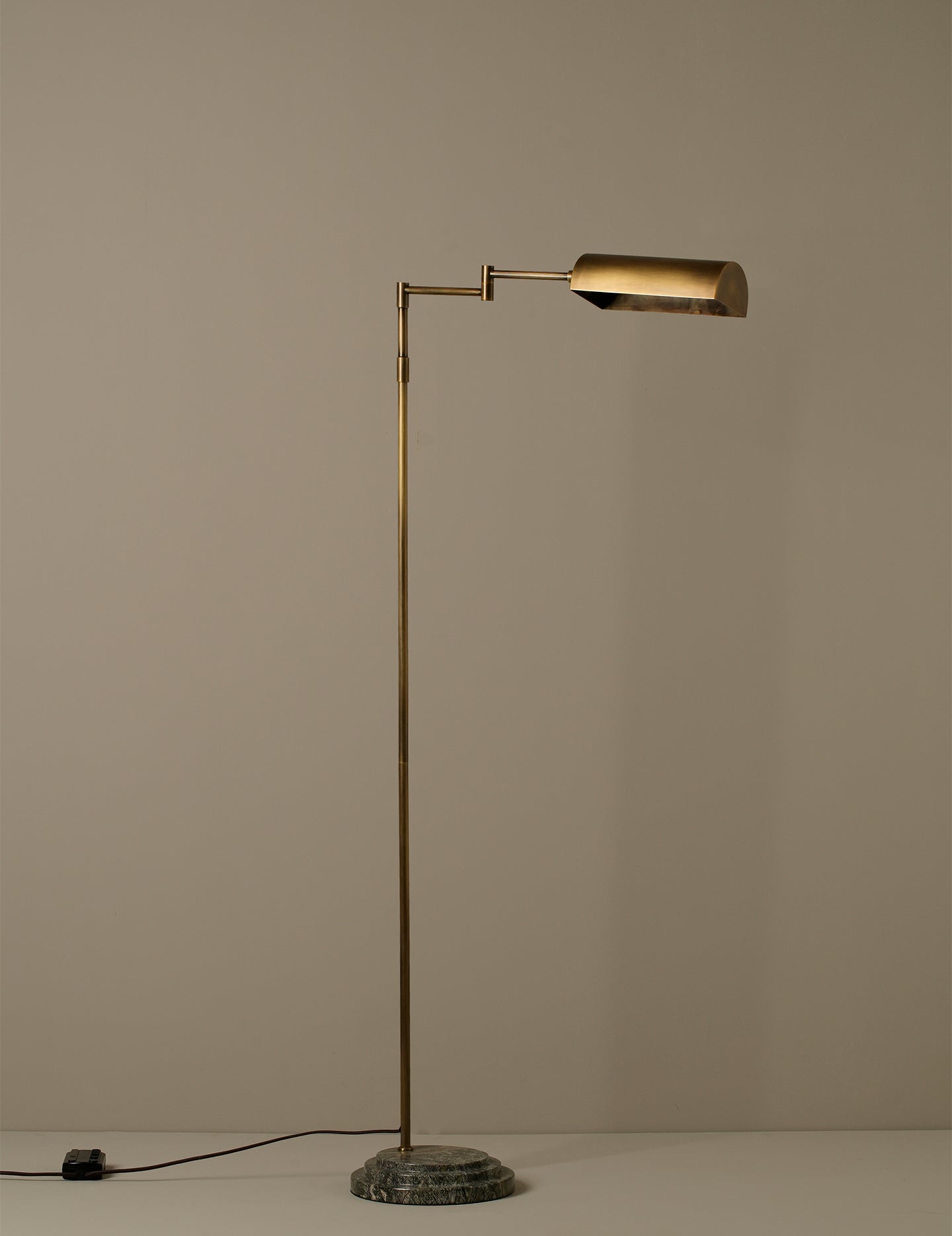 A modern floor lamp with a slim brass-colored metal stand, an adjustable arm, and a cylindrical shade, set on a round textured base against a plain beige background.