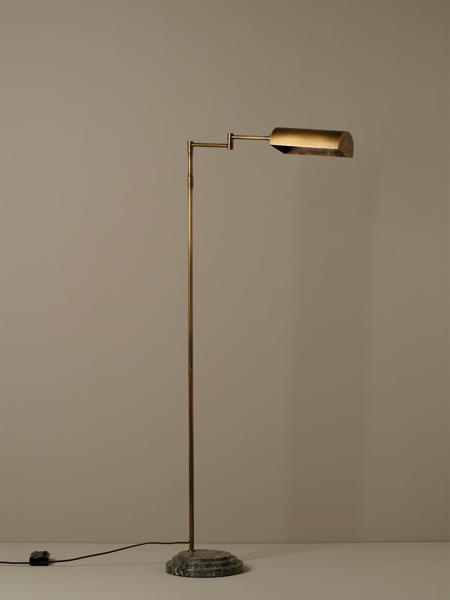 A modern floor lamp with a slim brass-colored metal stand, an adjustable arm, and a cylindrical shade, set on a round textured base against a plain beige background.