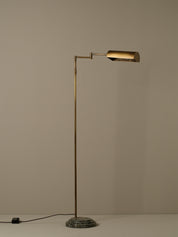 A modern floor lamp with a slim brass-colored metal stand, an adjustable arm, and a cylindrical shade, set on a round textured base against a plain beige background.