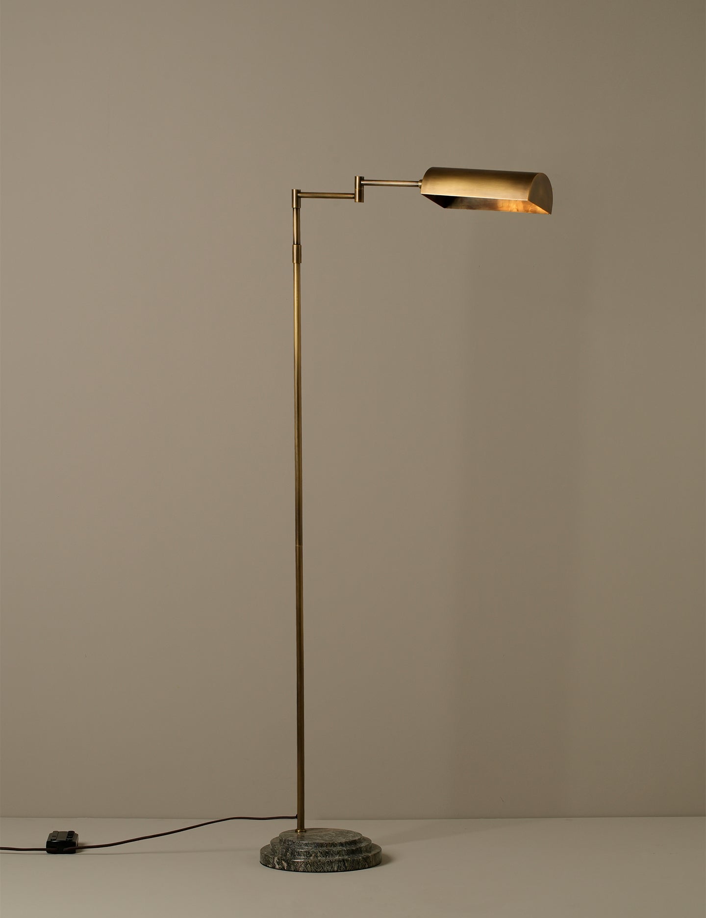 A modern floor lamp with a slim brass-colored metal stand, a cylindrical adjustable arm, and a matching cylindrical shade, set on a round textured base against a plain beige wall.