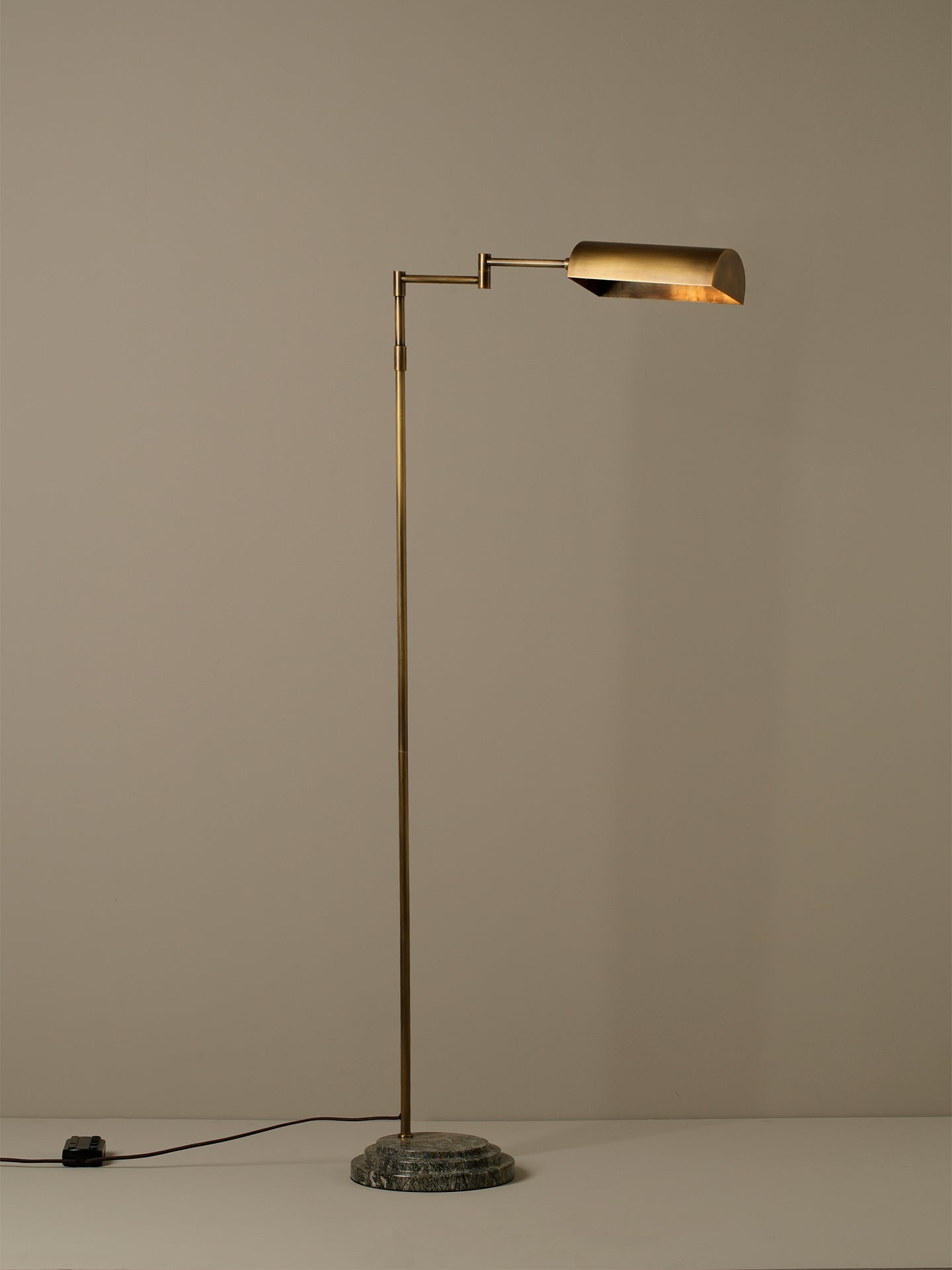 A modern floor lamp with a slim brass-colored metal stand, a cylindrical adjustable arm, and a matching cylindrical shade, set on a round textured base against a plain beige wall.