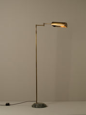 A modern floor lamp with a slim brass-colored metal stand, a cylindrical adjustable arm, and a matching cylindrical shade, set on a round textured base against a plain beige wall.