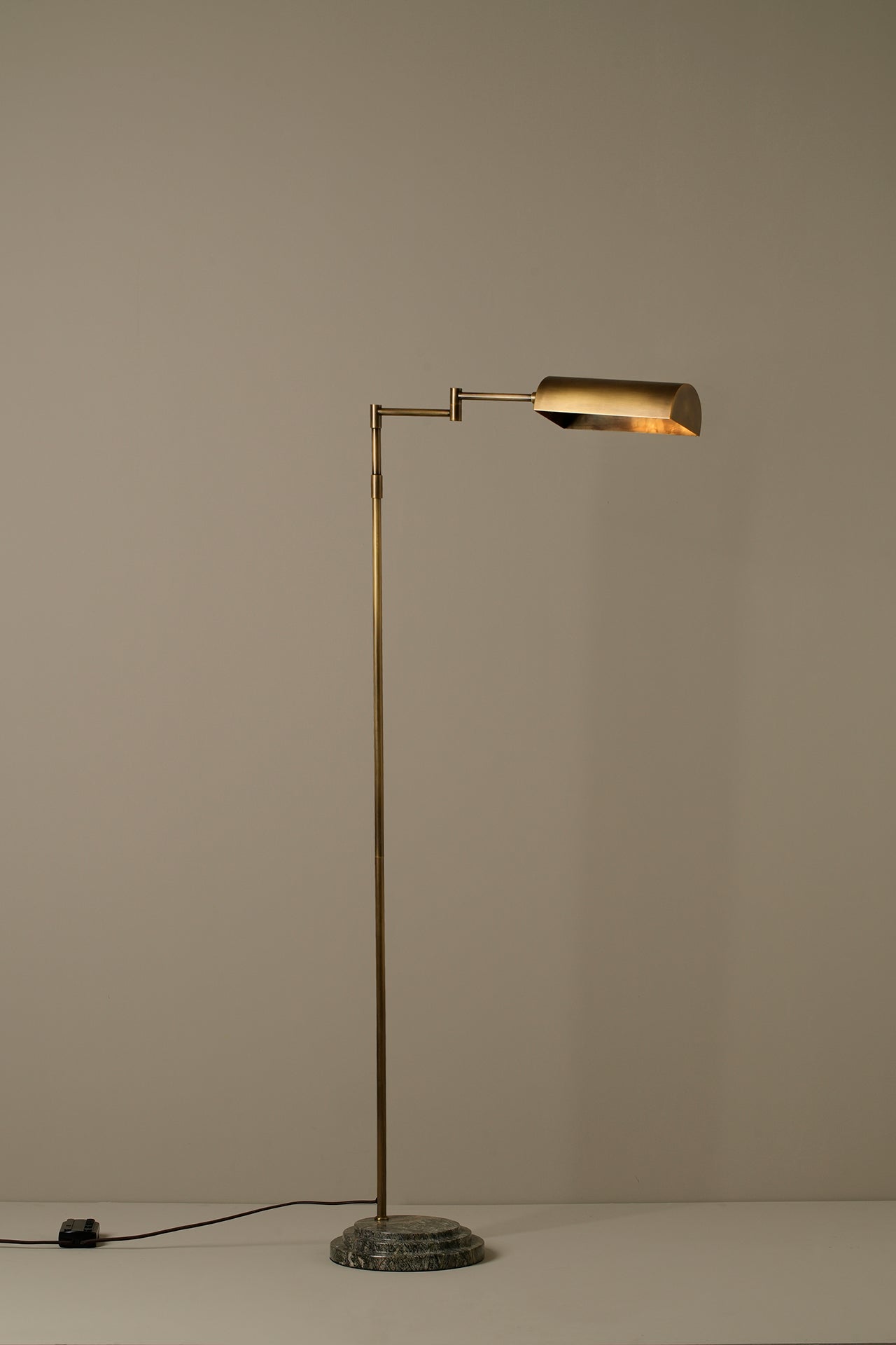 A modern floor lamp with a slim brass-colored metal stand and a rectangular shade, set against a plain beige wall with its power cord visible on the floor.
