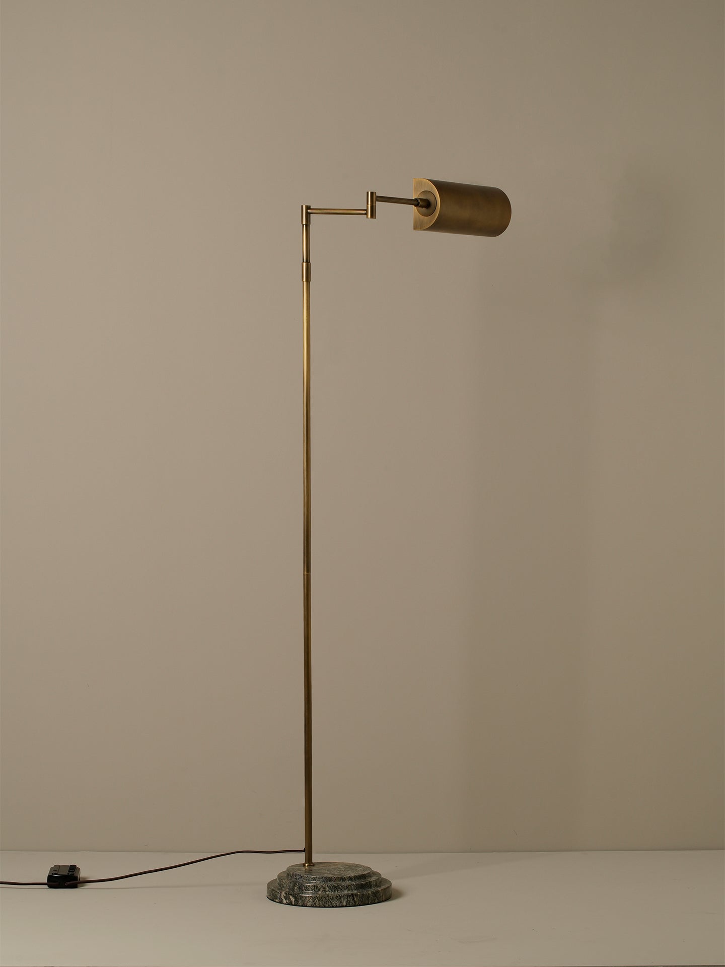 A mid-century modern floor lamp with a slim brass-colored metal pole, a cylindrical shade, and a round textured base, shown against a plain beige wall.