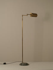 A mid-century modern floor lamp with a slim brass-colored metal pole, a cylindrical shade, and a round textured base, shown against a plain beige wall.