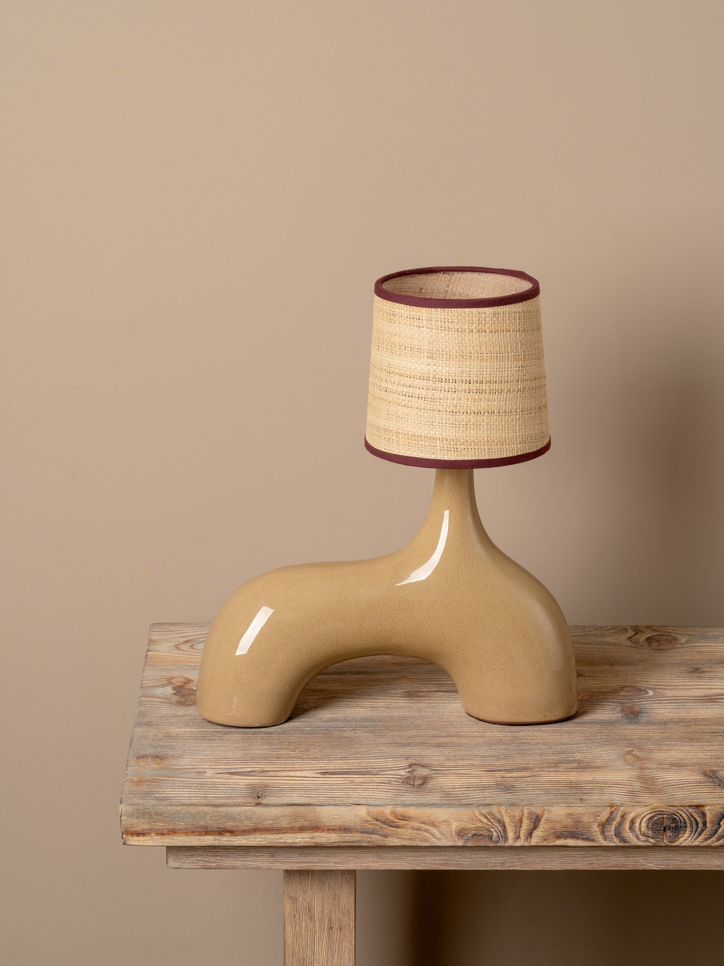 A sculptural ceramic table lamp with a glossy beige base and a textured beige and brown woven shade, displayed on a rustic wooden table against a solid beige background.