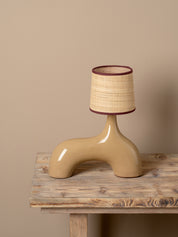A sculptural ceramic table lamp with a glossy beige base and a textured beige and brown woven shade, displayed on a rustic wooden table against a solid beige background.