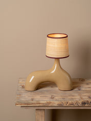 A sculptural table lamp with a glossy beige ceramic base and a textured beige woven shade trimmed in dark brown, displayed on a rustic wooden table against a solid beige background.