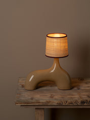 A sculptural table lamp with a glossy tan ceramic base and a textured beige woven shade, lit with a warm glow, displayed on a rustic wooden table against a solid taupe background.