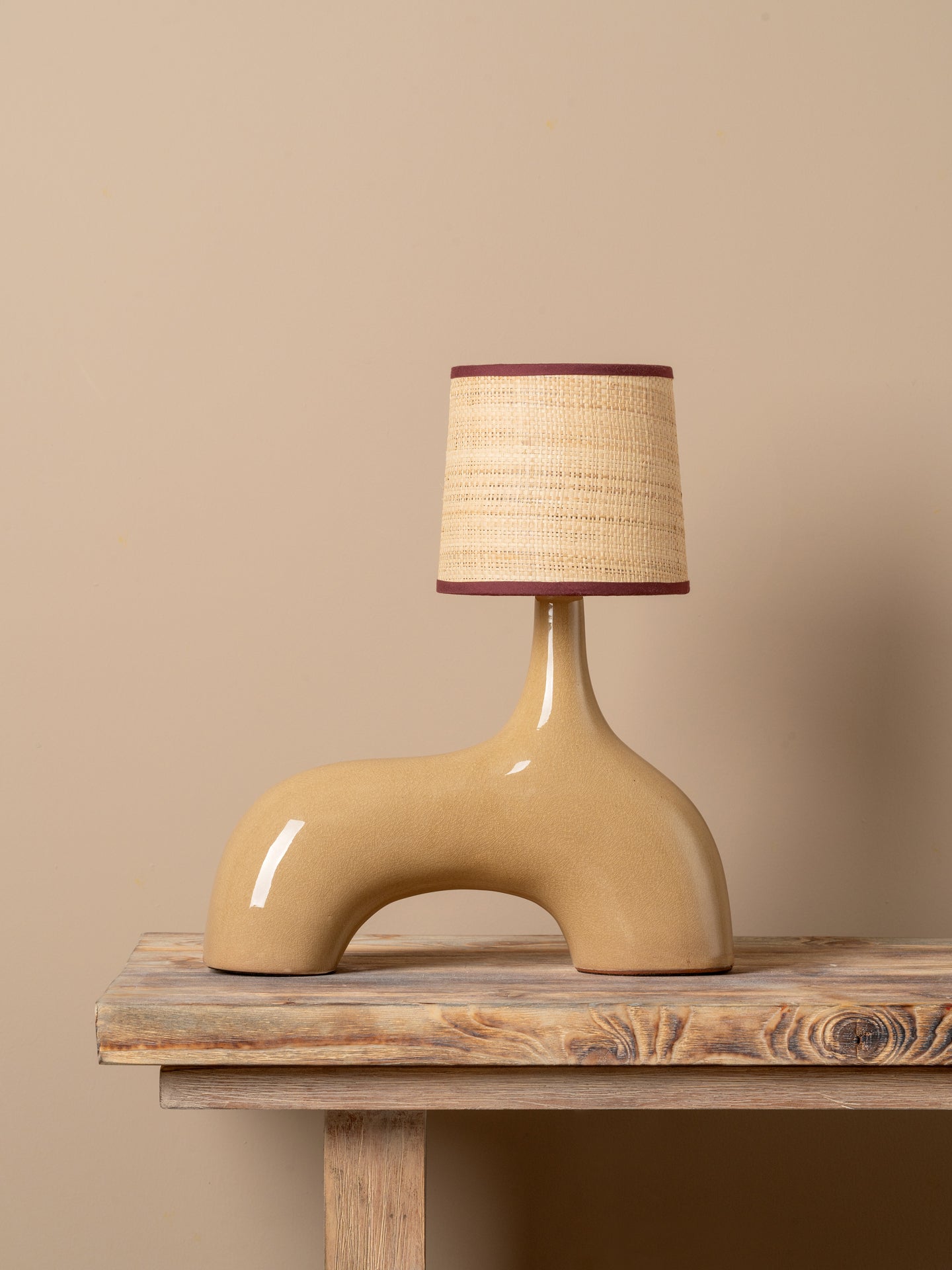 A sculptural ceramic table lamp with a glossy beige base and a woven straw-colored shade trimmed in dark red, displayed on a rustic wooden table against a solid beige background.