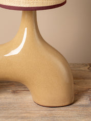 Close-up of a sculptural ceramic lamp base in a warm beige glaze, with a woven straw shade and dark burgundy trim visible at the top, set on a wooden surface against a beige background.