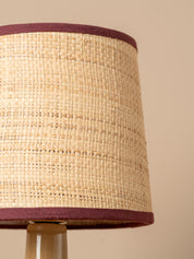 Close-up of a woven natural fiber lampshade with dark burgundy fabric trim, shown against a plain beige background.