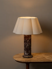 A table lamp with a cream pleated fabric shade and a dark brown marble base, illuminated and placed on a wooden side table against a warm gray wall.