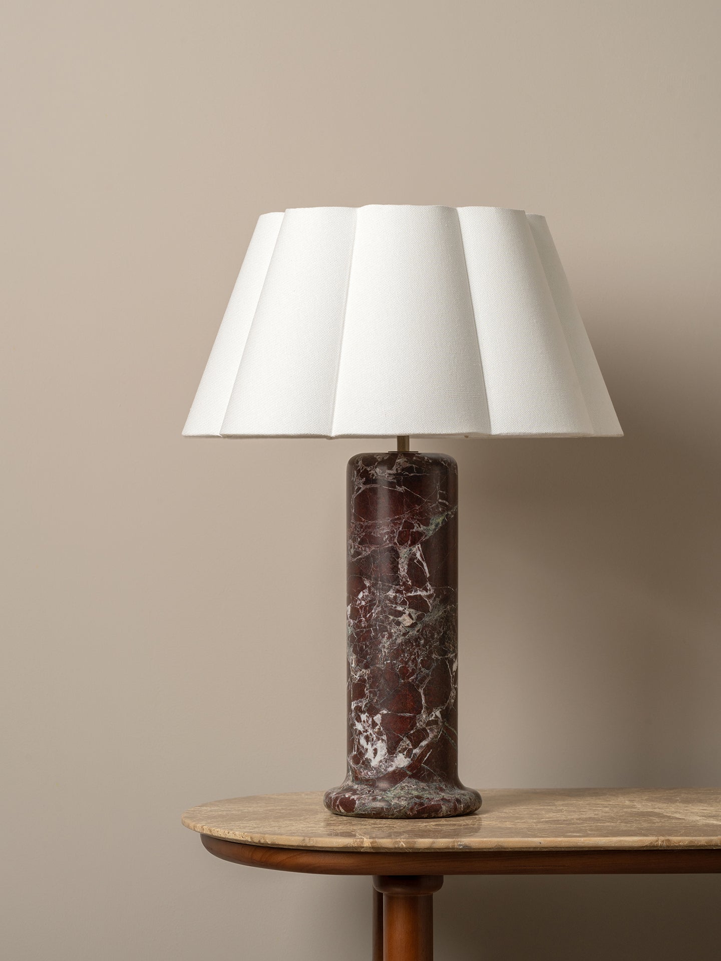 A table lamp with a white pleated fabric shade and a dark reddish-brown marble base, displayed on a wooden side table against a neutral beige wall.