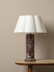 A table lamp with a white pleated fabric shade and a dark reddish-brown marble base, displayed on a wooden side table against a neutral beige wall.