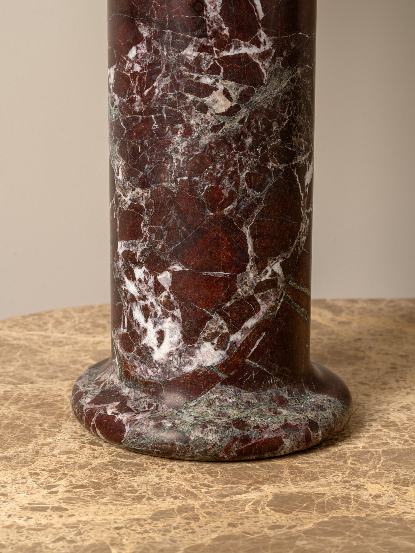 A close-up of a cylindrical pedestal or column made of polished dark red marble with prominent white and gray veining, standing on a beige stone surface against a neutral beige background.