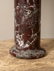 A close-up of a cylindrical pedestal or column made of polished dark red marble with prominent white and gray veining, standing on a beige stone surface against a neutral beige background.