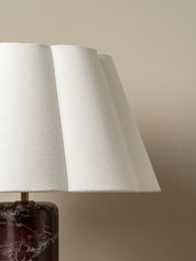 A close-up of a table lamp with a textured white pleated fabric shade and a dark brown marble base, set against a solid beige background.