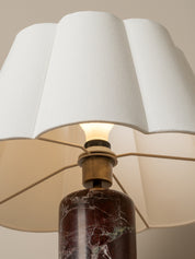 A close-up of a table lamp with a white pleated fabric shade, a brass metal collar, and a dark brown marble base with white veining, shown against a warm beige background.
