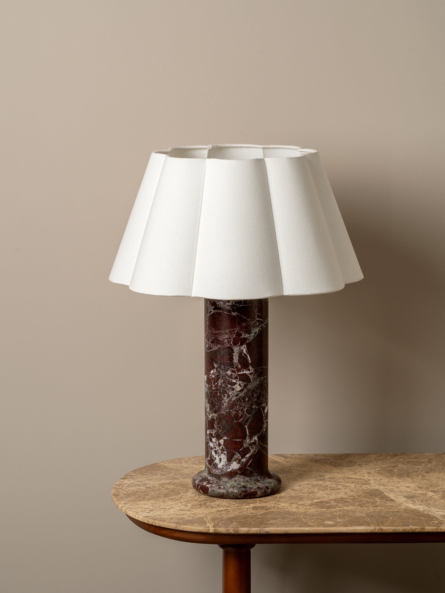 A table lamp with a white pleated fabric shade and a dark reddish-brown marble base, displayed on a round stone-topped side table against a warm beige wall.