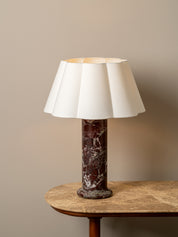 A table lamp with a white pleated fabric shade and a dark reddish-brown marble base, displayed on a wooden table against a neutral beige wall.