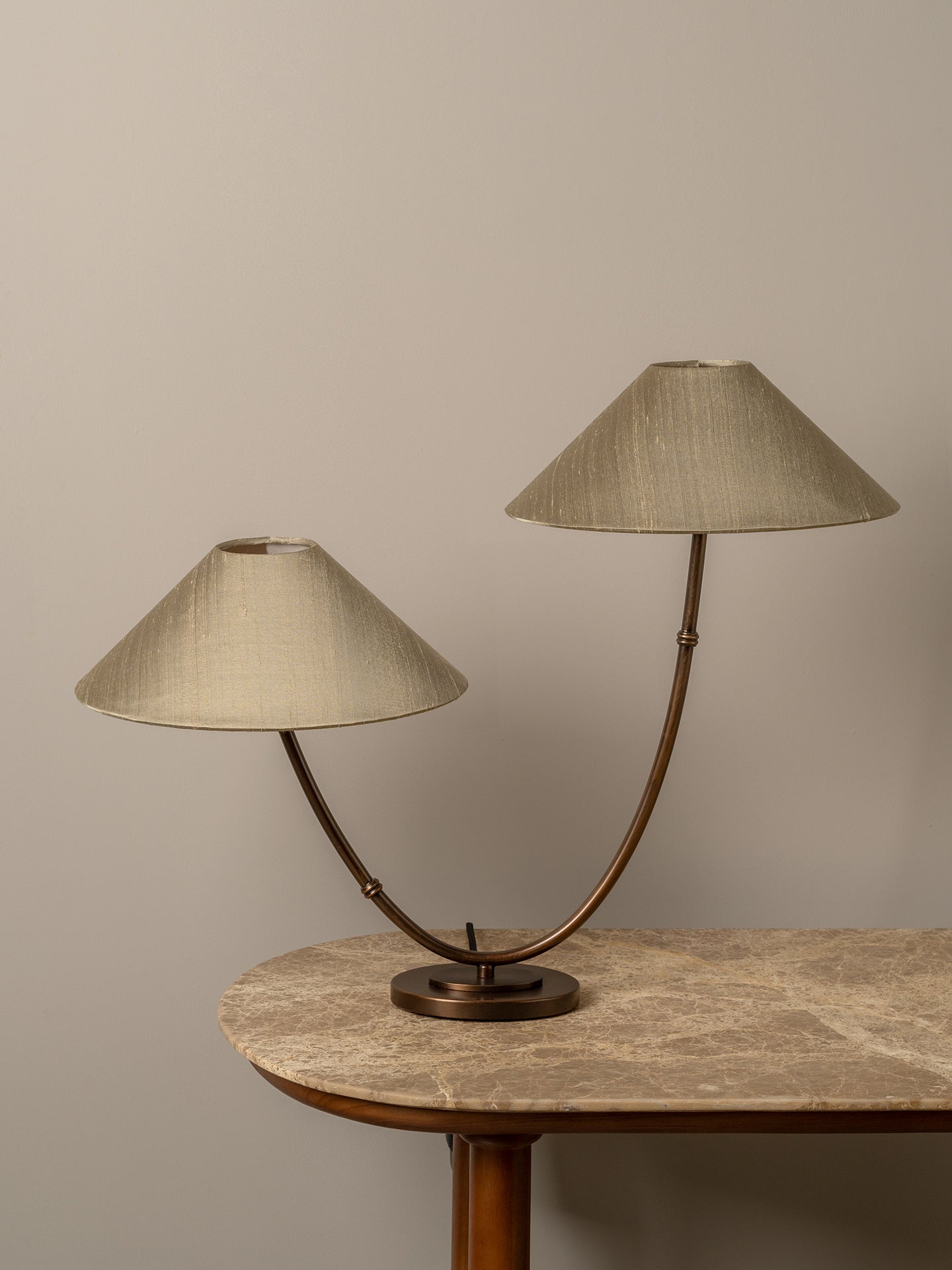A two-light table lamp with conical beige fabric shades and curved bronze metal arms, displayed on a stone-topped table against a neutral beige wall.