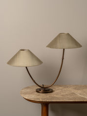 A two-light table lamp with conical beige fabric shades and curved bronze metal arms, displayed on a stone-topped table against a neutral beige wall.