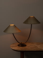 A two-light table lamp with conical fabric shades and curved dark metal arms, shown lit on a stone-topped table against a dark gray wall.