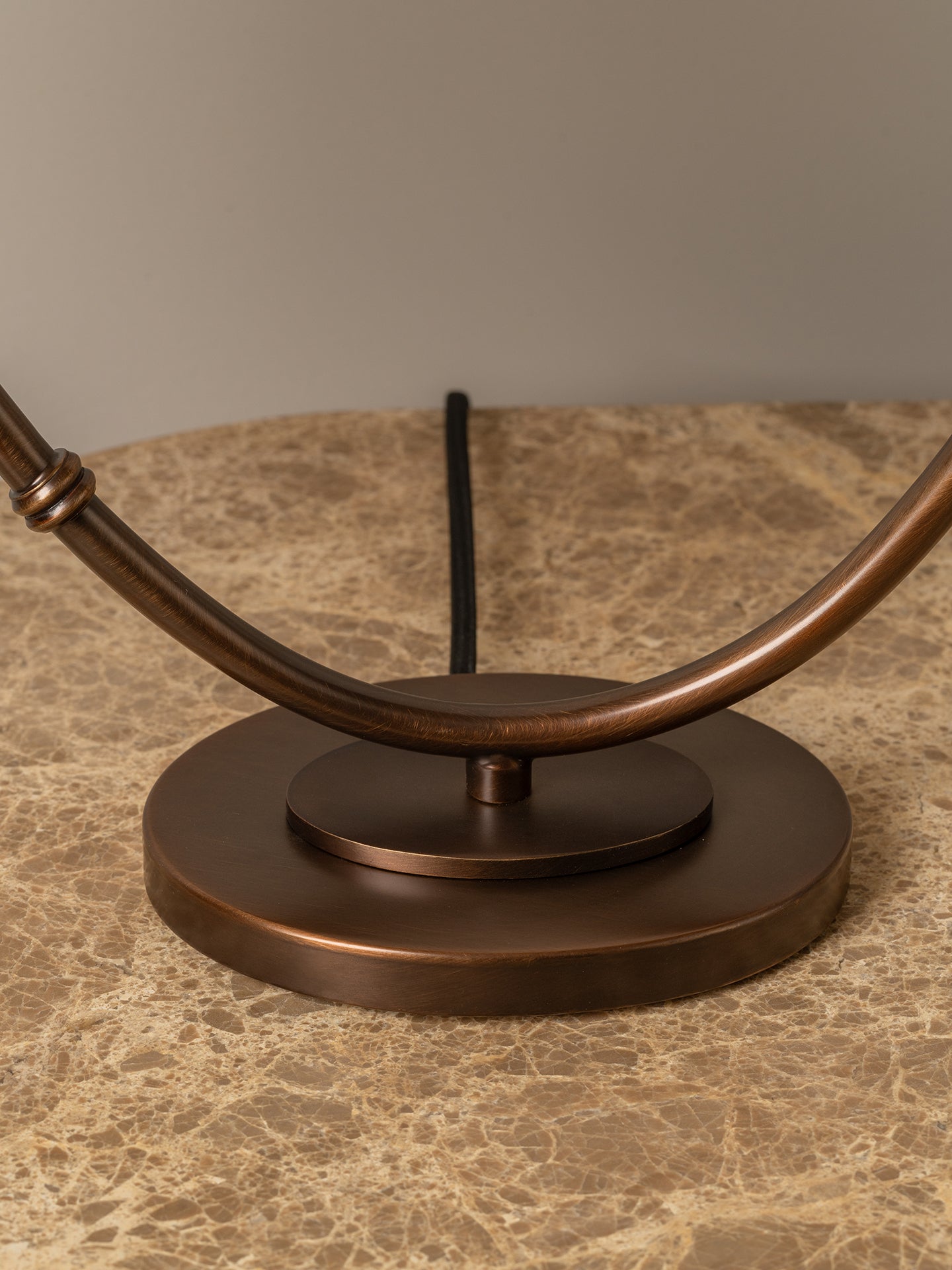 Close-up of a bronze metal table lamp base and curved arm on a beige marble surface against a neutral gray background.