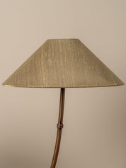 A close-up of a floor lamp with a tapered fabric shade in a textured beige-gold material and a curved metallic stem in a warm bronze finish, set against a plain beige background.