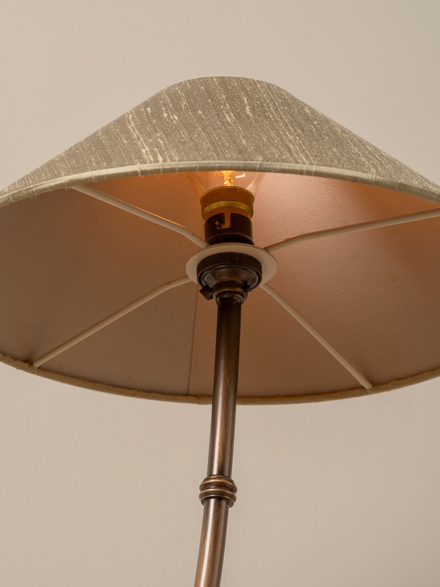 A close-up of a floor lamp with a textured beige fabric shade, a warm glowing light bulb, and a dark bronze metal pole against a neutral beige background.