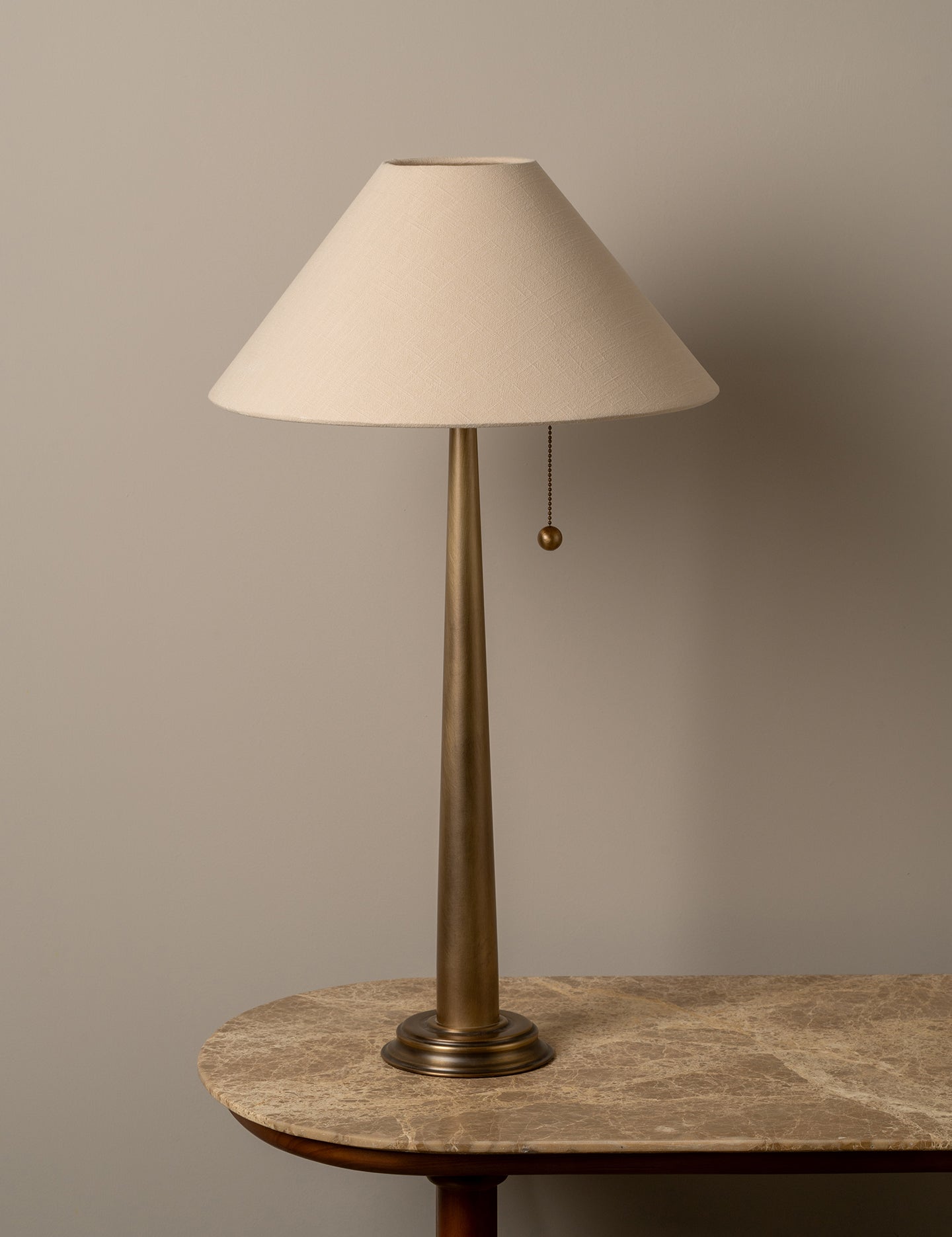 A table lamp with a tapered off-white fabric shade and a slim brass-colored metal base, shown resting on a stone-topped table against a neutral beige wall.
