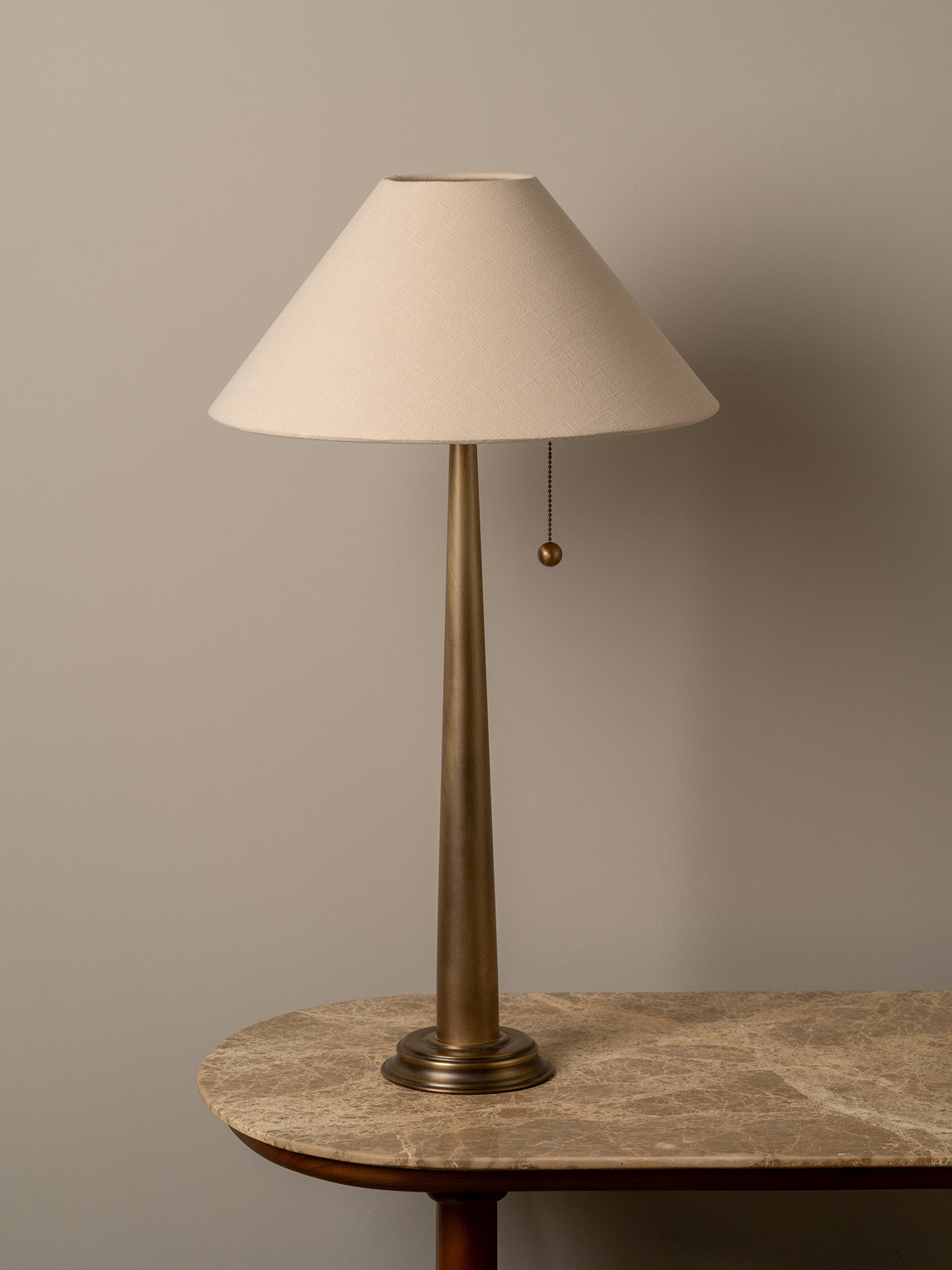 A table lamp with a tapered off-white fabric shade and a slim brass-colored metal base, shown resting on a stone-topped table against a neutral beige wall.