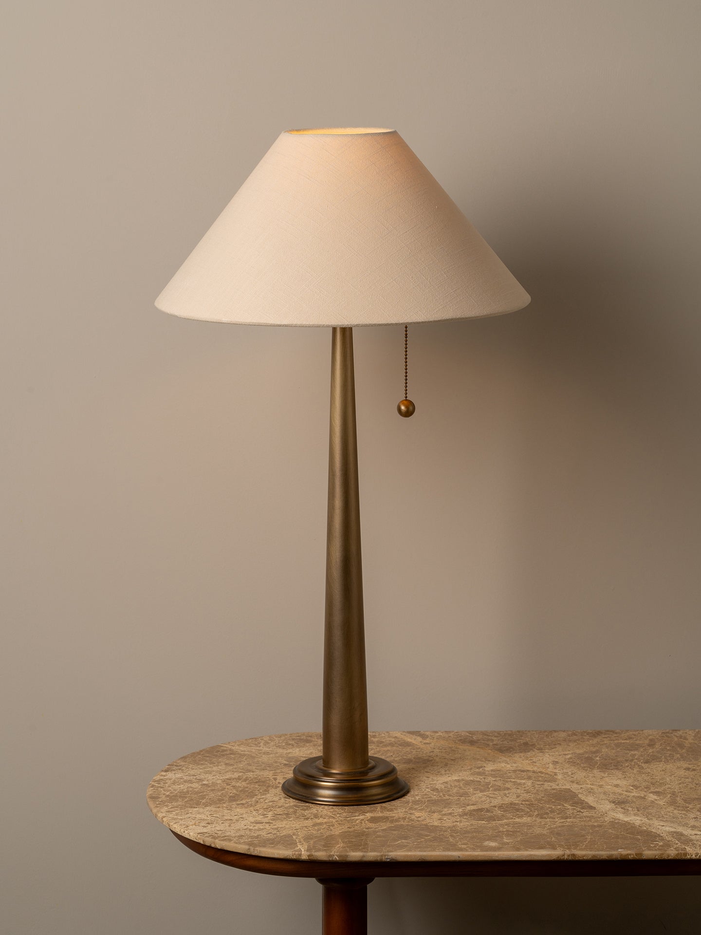 A table lamp with a tapered off-white fabric shade, a slim brass-toned metal stem, and a pull chain, displayed on a stone-topped table against a warm gray wall.