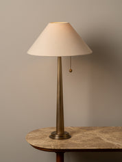 A table lamp with a tapered off-white fabric shade, a slim brass-toned metal stem, and a pull chain, displayed on a stone-topped table against a warm gray wall.