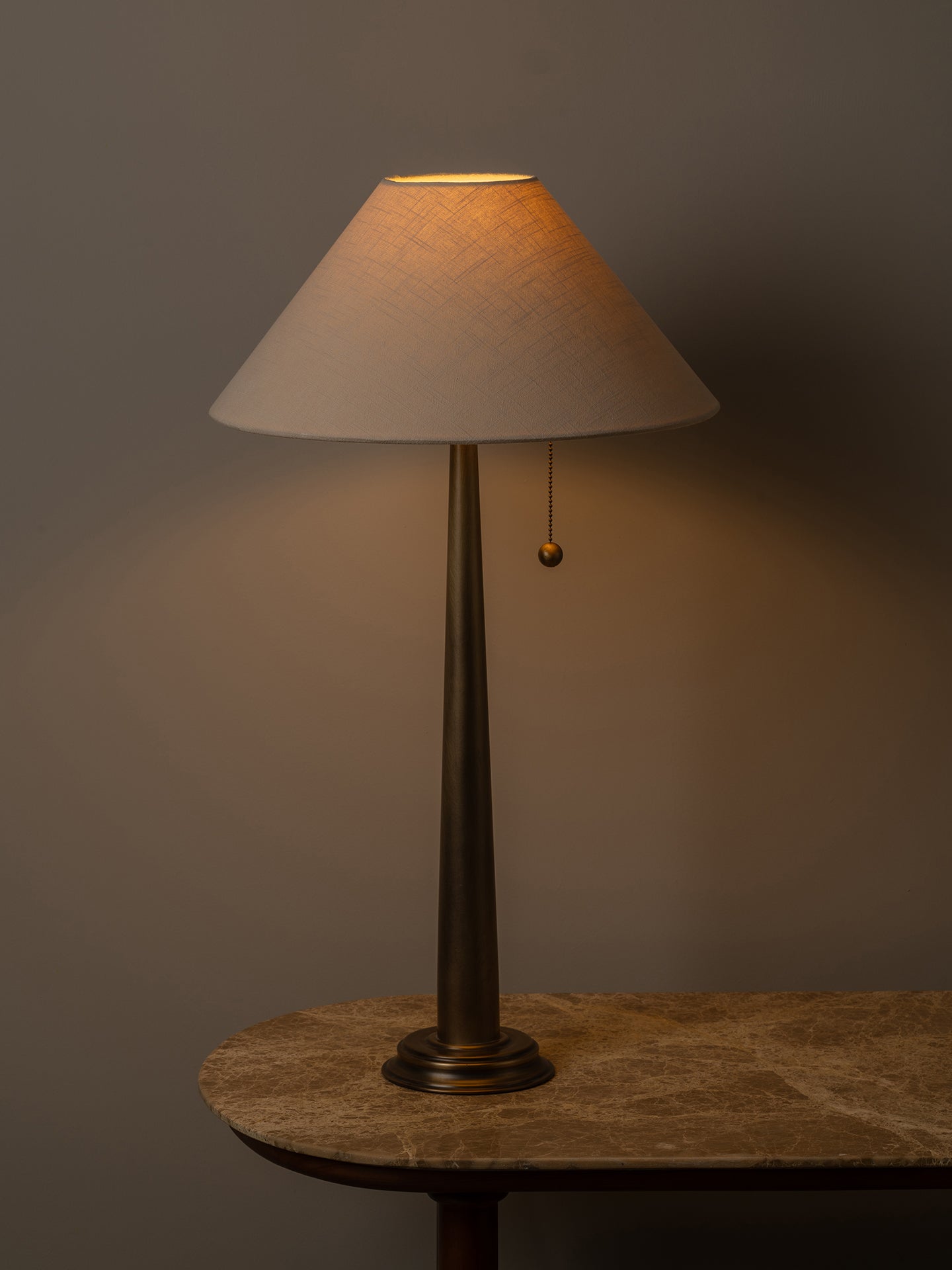 A table lamp with a tapered off-white fabric shade and a dark bronze metal stem, shown lit on a stone-topped table against a dark gray wall.