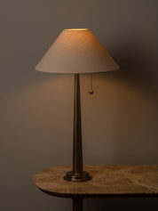 A table lamp with a tapered off-white fabric shade and a dark bronze metal stem, shown lit on a stone-topped table against a dark gray wall.