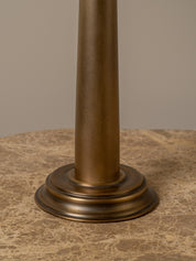 Close-up of a cylindrical column-style lamp base in a warm bronze finish, standing on a round marble-patterned surface against a plain beige background.