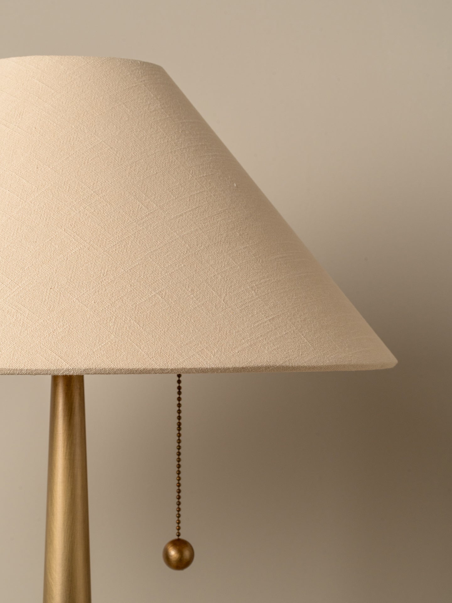 A close-up of a table lamp with a textured beige fabric shade and a brushed brass base, featuring a hanging pull chain with a round ball finial against a warm beige background.