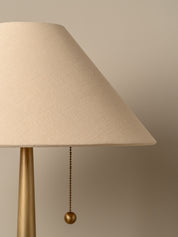 A close-up of a table lamp with a textured beige fabric shade and a brushed brass base, featuring a hanging pull chain with a round ball finial against a warm beige background.