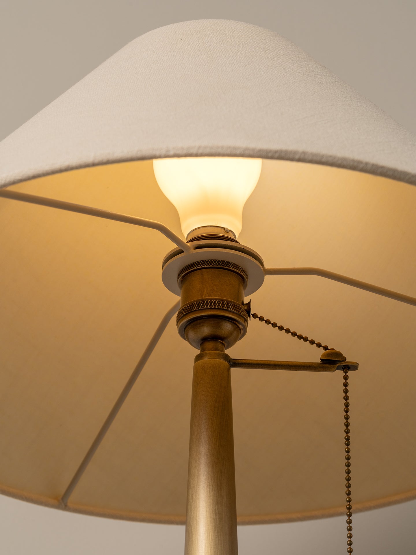 Close-up of a floor lamp with a large off-white fabric drum shade, a warm glowing light bulb, and a brass-toned metal stem and pull chain against a neutral background.