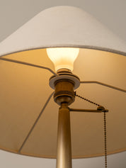 Close-up of a floor lamp with a large off-white fabric drum shade, a warm glowing light bulb, and a brass-toned metal stem and pull chain against a neutral background.