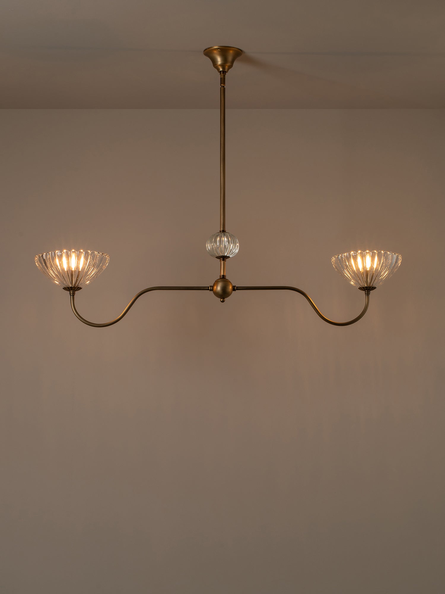 A two-light brass chandelier with fluted clear glass shades, suspended from a central clear glass bead, shown against a plain beige wall.