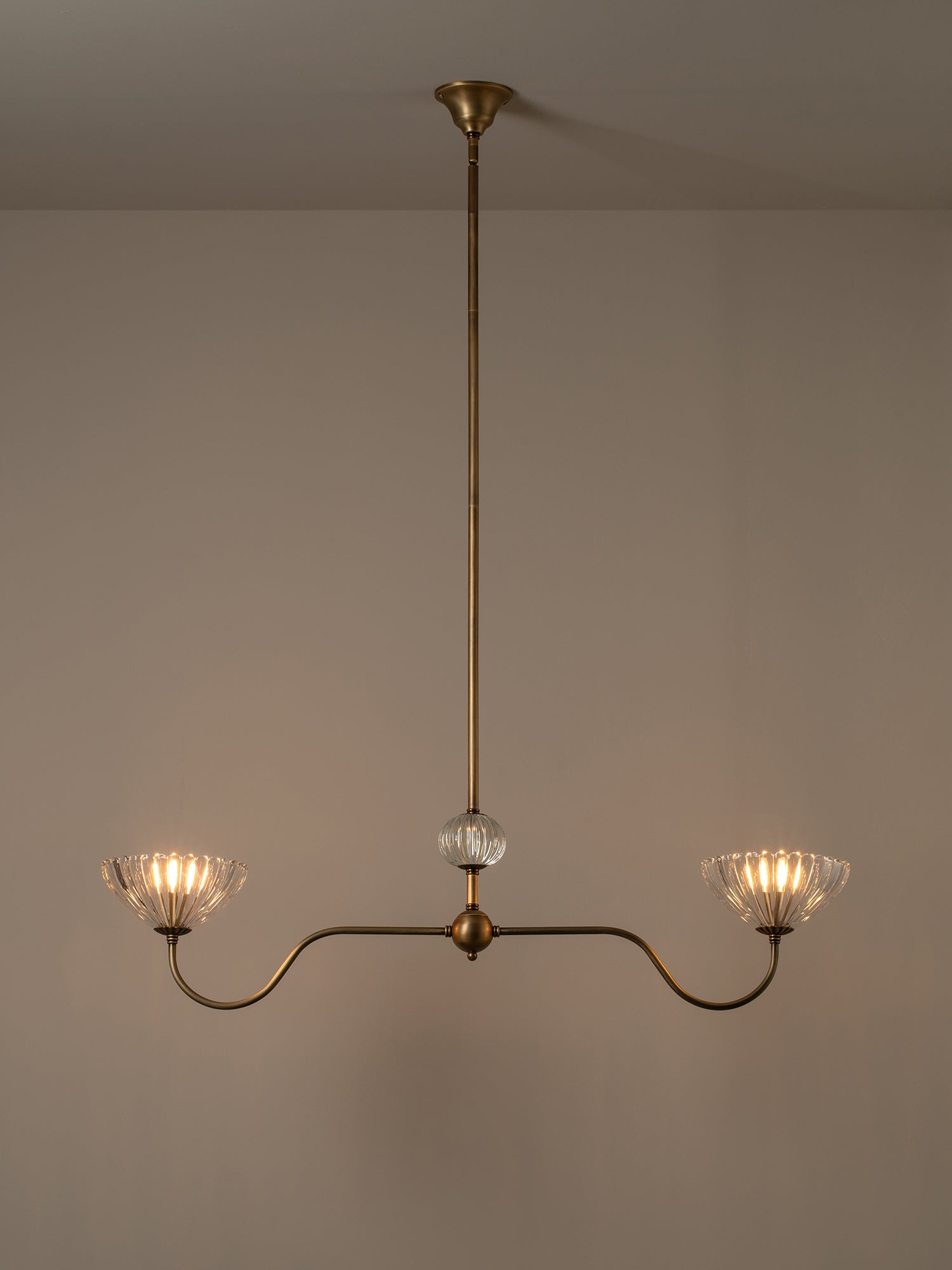 A two-light brass chandelier with curved arms, clear ribbed glass shades, and a central clear glass sphere, shown hanging against a plain beige wall.