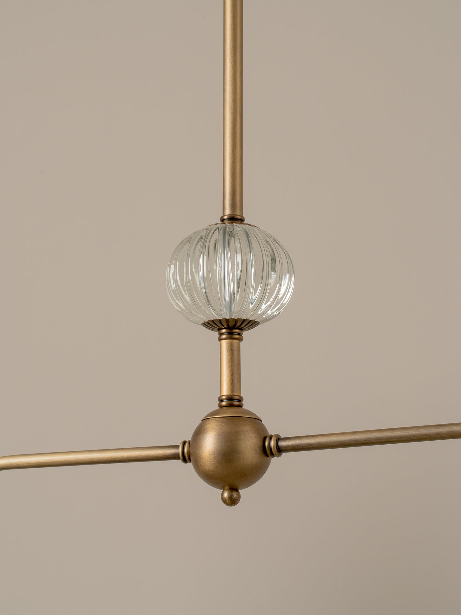 A close-up of a modern pendant light fixture with a brushed brass finish and a ribbed clear glass globe shade, shown against a solid beige background.