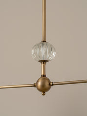 A close-up of a modern pendant light fixture with a brushed brass finish and a ribbed clear glass globe shade, shown against a solid beige background.