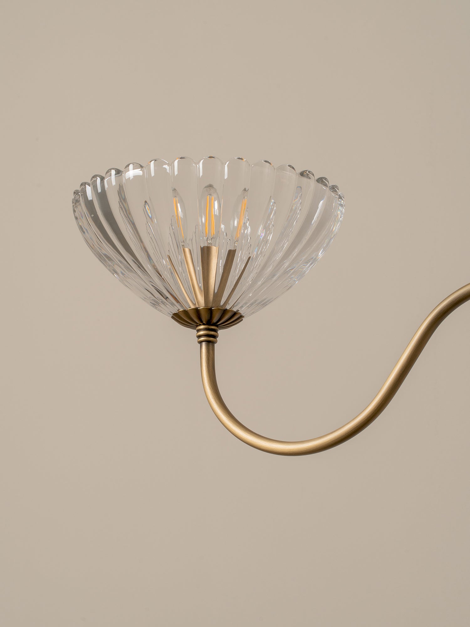 A close-up of a wall or ceiling light fixture with a clear ribbed glass shade and a curved brass or gold metal arm against a warm beige background.