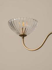 A close-up of a wall or ceiling light fixture with a clear ribbed glass shade and a curved brass or gold metal arm against a warm beige background.